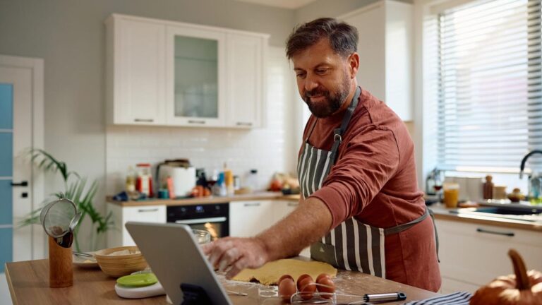 A man in an apron in a kitchen smiles, reaching toward a smart tablet.