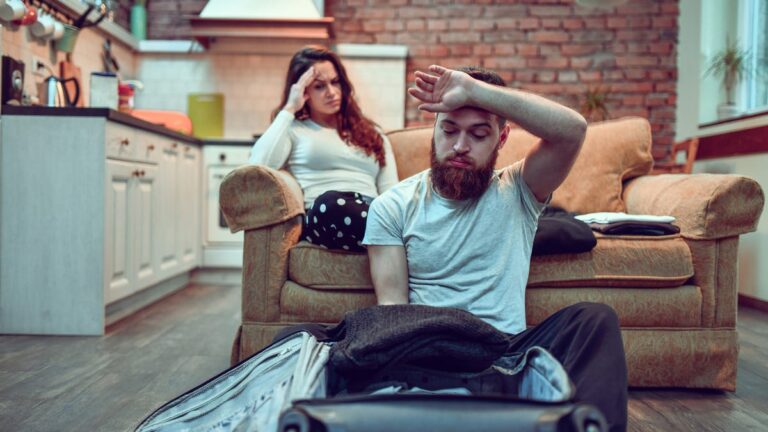 A couple frowns over an open suitcase against a sofa and brick wall.