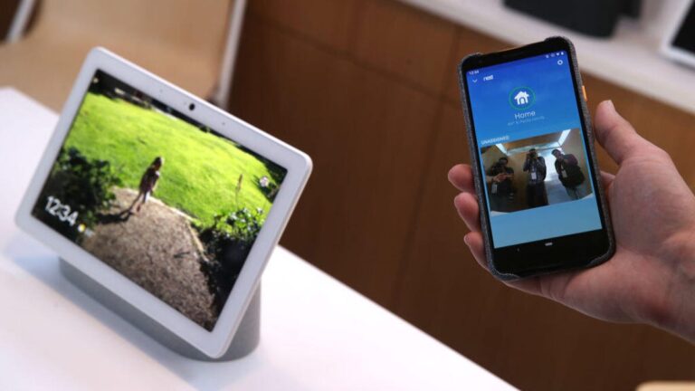 A hand holds a phone showing a Nest cam view beside a Nest display on a white counter.