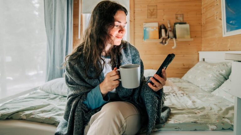 A woman in a robe sits on a bed with a cup and looks at her phone.