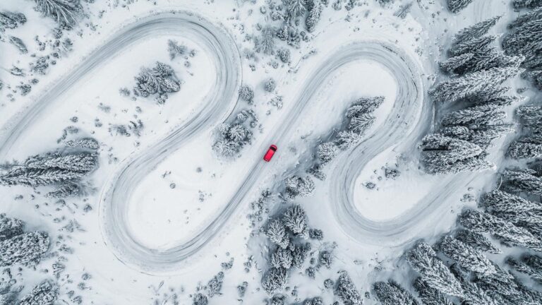 An aerial photo of a red car driving along a snowy road surrounded by trees
