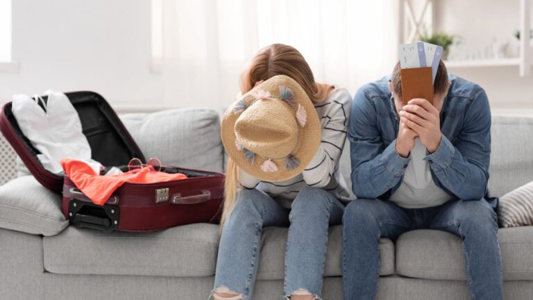 Two people sit on a sofa by a suitcase, hiding their faces with a hat and tickets.