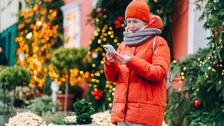 Woman in read outside holiday-decorated homes looks at her phone.