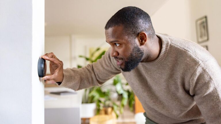A man leans over to adjust a thermostat dial on a home wall.