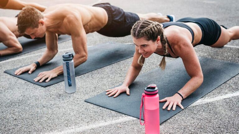 An Expert-Approved Strength Training and Cardio Routine That Will Get You Fit A young man and woman planking outside over their yoga mats with their water bottles in the foreground.